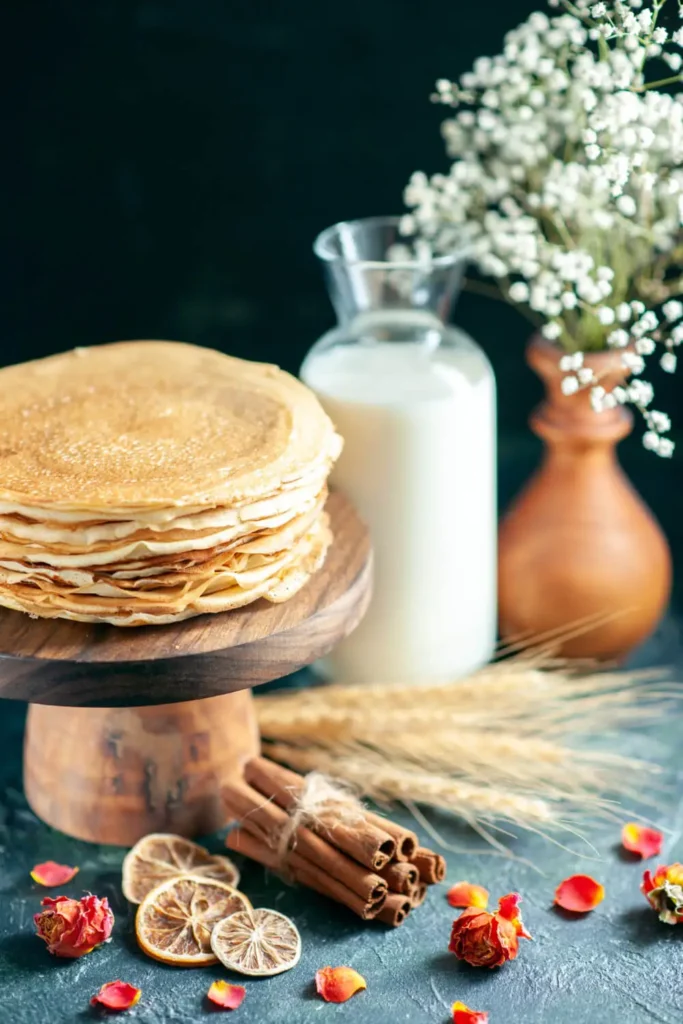 A stack of golden pancakes on a wooden stand with milk and floral decoration in the background